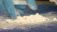 Kokainpulver liegt auf einem Tisch und eine Hand mit blauen Handschuhen fasst dieses an © Screenshot 