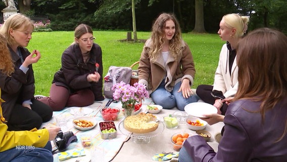 Vier junge Frauen sitzen auf einer Picknick-Decke bei Kuchen und anderen Leckereien. © Screenshot 