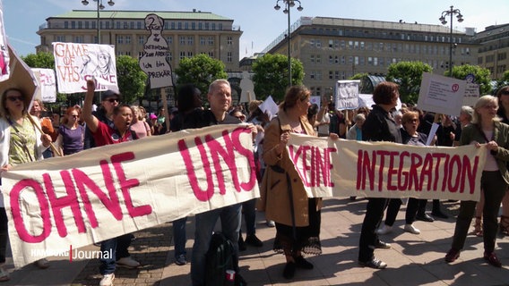 Menschen halten auf dem Hamburger Rathausmarkt ein Banner hoch. Darauf steht "Ohne uns keine Integration". © Screenshot 