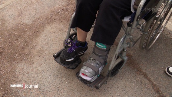 Nahaufnahme von den Beinen einer Person, die im Rollstuhl sitzt. Der linke Fuß ist in einer Schiene. © Screenshot 