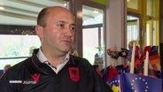 Albert Bisaku im Interview. Er trägt ein albanisches Nationaltrikot. Im Hintergrund stehen diverse Flaggen von vielen Ländern, die an der EM teilnehmen. © Screenshot 