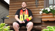Ein Mann mit Gipsarm in Deutschlandfarben sitzt vor seiner Hütte. © Screenshot 