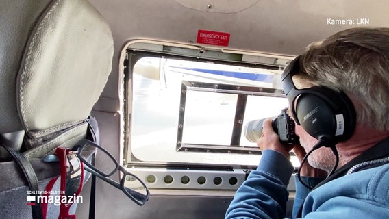 Ein Mann fotografiert mit einer Kamera aus einem fliegenden Hubschrauber raus. Er trägt Ohrschützer. © Screenshot 