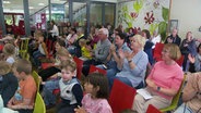 Kinder, Eltern und Großeltern sitzen applaudierend im Publikum bei einer Aufführung in einem Schulgebäude. © Screenshot 