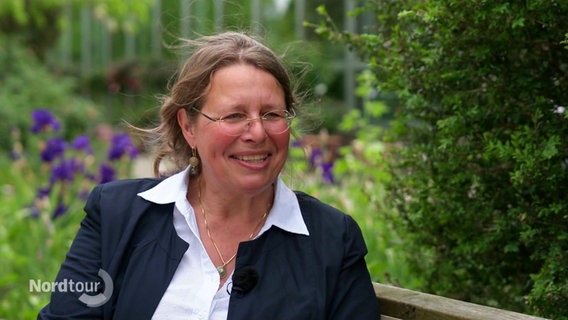 Prof. Dr. Anke Seegert sitzt auf einer Bank im Grünen und lacht in die Kamera. © Screenshot 