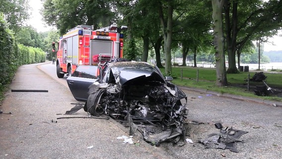 Ein stark zerstörtes Auto steht am Straßenrand, im Hintergrund ein Einsatzwagen der Feuerwehr. © Screenshot 