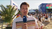 Reporter Sebastian Rieck am 23.06.2024 live am Heiligengeistfeld. Hinter ihm sitzen Menschen auf Stühlen, der Boden ist mit weißem Sand bedeckt und die Sonne scheint. © Screenshot 