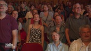 Das Publikum eines Konzerthauses erhebt sich zum gemeinsamen Singen von den Plätzen. © Screenshot 