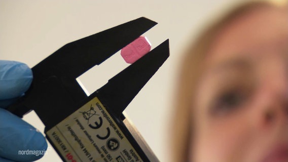 Eine Frau hält eine Messzange, in der eine rosafarbene Extasy-Pille klemmt. © Screenshot 