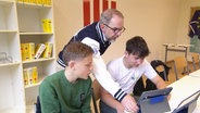Ein Lehrer und zwei Schüler schauen auf ein Tabletcomputer. © Screenshot 