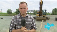NDR Reporter Thees Jagels steht vor einem Bagger auf einer Baustelle. © Screenshot 