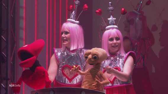 Zwei als Außerirdische verkleidete Frauen mit Puppen auf einer Bühne. © Screenshot 