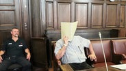 Ein Angeklagter sitz auf der Anklagebank im Oberlandesgericht Hamburg und hält sich eine Akte vor sein Gesicht. Neben dem Angeklagten sitz ein Mitarbeiter der Justiz. © Screenshot 