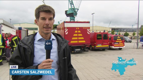 NDR Reporter Carsten Salzwedel berichtet vor Ort. Hinter ihm sind Feuerwehrkräfte und Einsatzfahrzeuge zu sehen. © Screenshot 