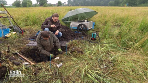 Forscher bei der Entnahme von Bodenproben im Moor. Sie stehen auf einer Wiese in einer ausgehobenen Grube. Das Gras ist hoch, der Boden torfig. Es regnet, ein kleiner Wagen mit Messutensilien ist mit einem Regenschirm abgedeckt. © Screenshot 