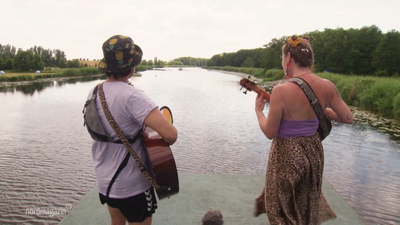 Zwei Personen mit Gitarren stehen mit dem Rücken zur Kamera auf dem Dach eines Floßes und musizieren. Vor ihnen erstreckt sich eine Flusslandschaft. © Screenshot 