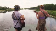 Zwei Personen mit Gitarren stehen mit dem Rücken zur Kamera auf dem Dach eines Floßes und musizieren. Vor ihnen erstreckt sich eine Flusslandschaft. © Screenshot 