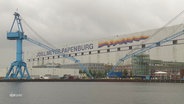 Blick auf das Gelände der Meyer-Werft in Papenburg. Direkt vor der großen Halle steht ein blauer Kran. © Screenshot 