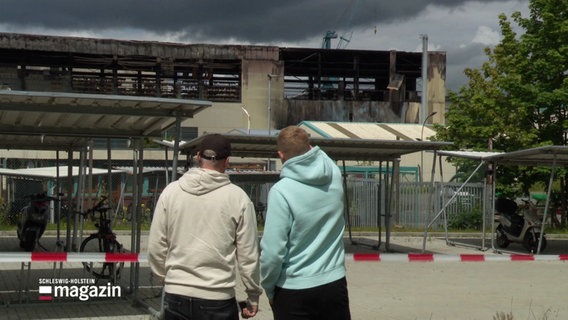 Zwei Personen vor einer ausgebrannten Lagerhalle. © Screenshot 