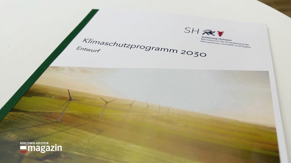 Auf einem weißen Tisch liegt ein Heft mit der Überschrift "Klimaschutzprogramm 2023. Entwurf". Darunter ist ein Bild von einer grünen Landschaft mit Windrädern, darüber strahlt gelbes Sonnenlicht. © Screenshot 