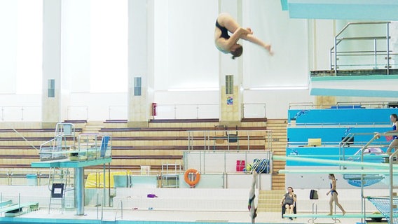 Ein Mädchen springt in einer Schwimmhalle von einem Turm. © Screenshot 