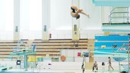 Ein Mädchen springt in einer Schwimmhalle von einem Turm. © Screenshot 