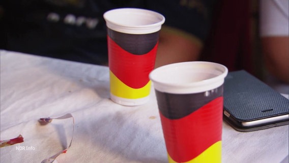 Zwei Plastik-Becher in den Farben der deutschen Flagge stehen auf einem Tisch. © Screenshot 