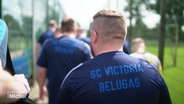 Ein Mann trägt ein dunkelblaues Fußballtrikot, auf dessen Rückseite der Schriftzug "SC Victoria Belugas" gedruckt ist. © Screenshot 