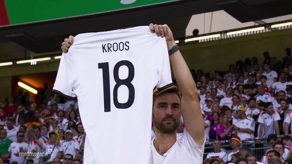 Ein Fußballfan hält ein Trikot mit der Nummer 18 und dem Namen Kroos hoch. © Screenshot 