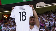 Ein Fußballfan hält ein Trikot mit der Nummer 18 und dem Namen Kroos hoch. © Screenshot 