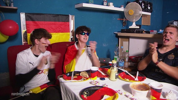 An einem mit Deutschland-Farben dekorierten Tisch jubeln drei Fußballfans. © Screenshot 