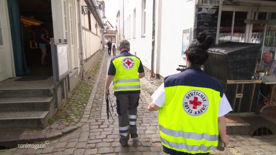 Minarbeiter des Deutschen Roten Kreuzes gehen in geben Westen durch eine Gasse. © Screenshot 