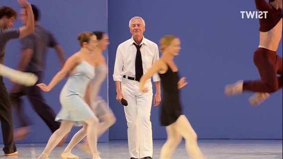 Choreograf John Neumeier geht langsam durch eine Gruppe tanzender Menschen. © Screenshot 