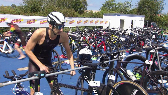 Ein Triathlon-Teilnehmer greift sich aus einer Reihe an Rennrädern seines heraus. © Screenshot 