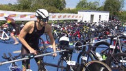 Ein Triathlon-Teilnehmer greift sich aus einer Reihe an Rennrädern seines heraus. © Screenshot 