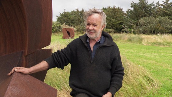 Der Künstler Jörg Plickat steht neben einer seiner Skulpturen aus Metall. © Screenshot 