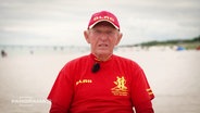 Rettungsschwimmer Klaus Scheler sitzt mit roter Cap und rotem T-Shirt am Strand und gibt ein Interview. © Screenshot 