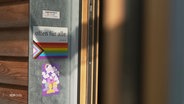 Ein Aufkleber auf einer Tür, darauf die Worte: "Offen für alle" und eine Regenbogenfahne. © Screenshot 