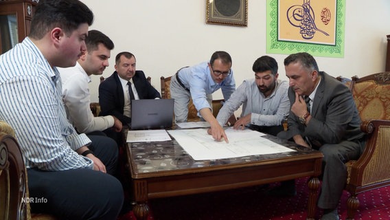 Eine Gruppe Männer arbeitet an einem Bauplan. © Screenshot 