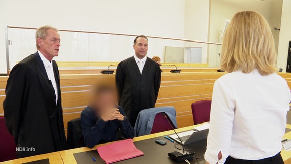 Der Angeklagte sitzt zwischen zwei Männern in schwarzen Roben vor Gericht. © Screenshot 