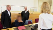 Der Angeklagte sitzt zwischen zwei Männern in schwarzen Roben vor Gericht. © Screenshot 