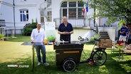 Dave Hänsel und der italienische Sprachlehrer Giuseppe kochen mit einer mobilen Küche auf einem Lastenfahrrad im Garten des italienischen Kulturinstitutes. © Screenshot 