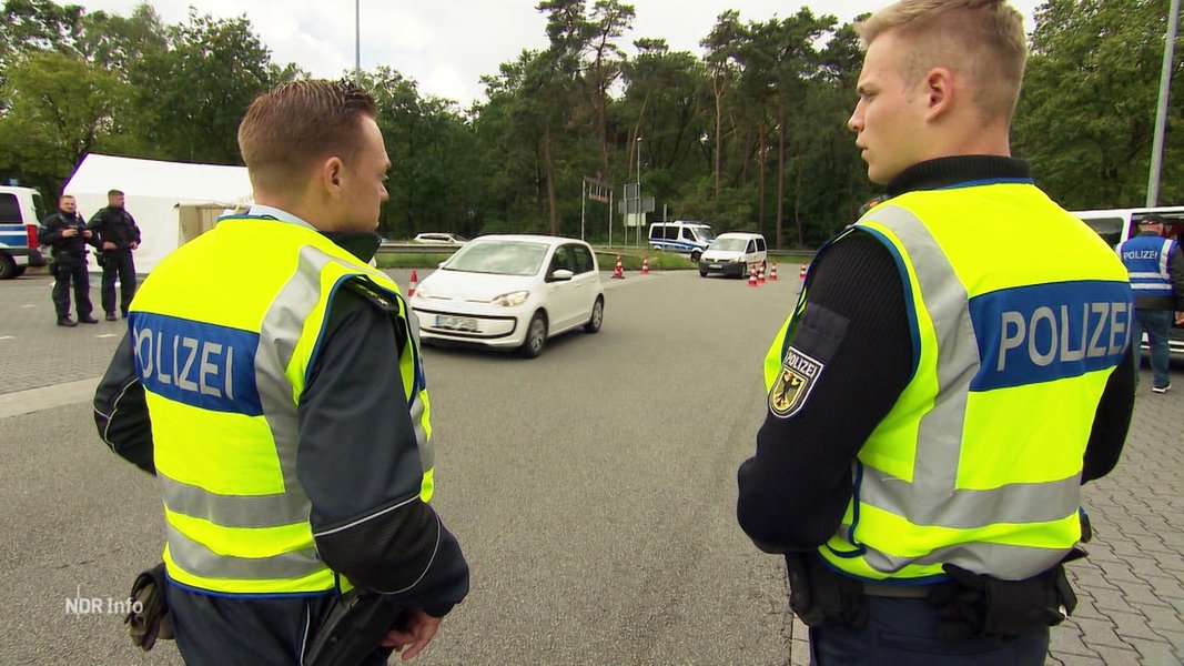 Zwei Polizisten an einem Parkplatz, in gelben Warnwesten und von schräg hinten gefilmt.