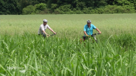 Zwei Personen fahren mit dem Fahrrad durch sehr hohes Gras. © Screenshot 