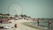 Ein sommerliche Tag in der Lübecker Bucht. Menschen tummeln sich auf dem Sand und im Wasser. Im Hintergrund: ein Riesenrad. © Screenshot 