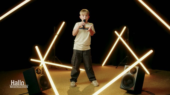 Der 10-Jährige Bjarne sing und schaut dabei in die Kamera. Er trägt einen blonden Topfschnitt. Um ihn herum sind helle Leuchtröhren verteilt, die dem Bild eine warme Ausstrahlung verleihen. © Screenshot 