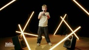 Der 10-Jährige Bjarne sing und schaut dabei in die Kamera. Er trägt einen blonden Topfschnitt. Um ihn herum sind helle Leuchtröhren verteilt, die dem Bild eine warme Ausstrahlung verleihen. © Screenshot 