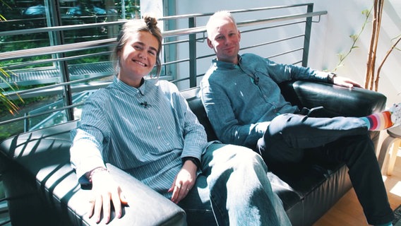 Moderatorin Theresa Hebert und Moderator Nils Söhrens sitzen neben Palmen auf einer Couch. © Screenshot 