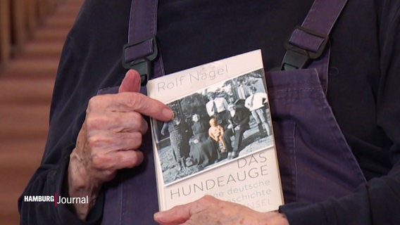 Alte Männerhände halten ein Buch und deuten auf das Titelbild, das ein schwarz-weiß Foto von ein paar Menschen zeigt, von denen ein Kind in Farbe dargestellt ist. © Screenshot 