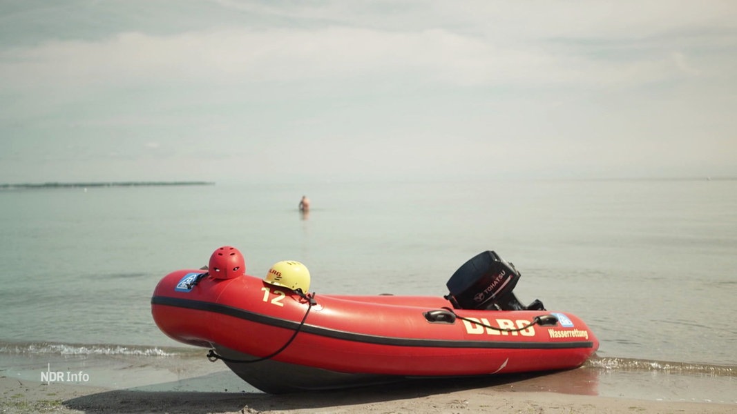 Ein rotes Rettungsboot der DLRG liegt an einem Strand. Im Hintergrund ist ein Badegast zu sehen.